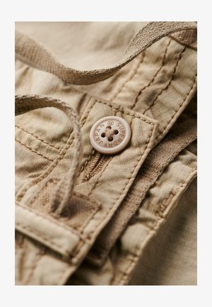 Beige stof tæt på med en tekstureret overflade, en rund knap mærket "SUPER JAPAN" og syede detaljer langs kanterne.