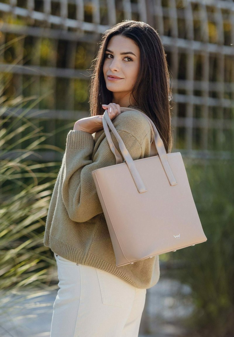 Hellbeige Tragetasche mit zwei oberen Tragegriffen, rechteckiger Form, glatter Textur und einem Logo auf der Vorderseite. Kombiniert mit einem beigen Pullover und weißen Hosen.