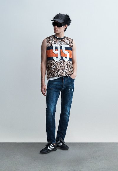 Homme portant une chemise sans manches à imprimé léopard avec le chiffre "95", un jean bleu, une casquette noire et de grandes lunettes de soleil noires, debout devant un fond uni.