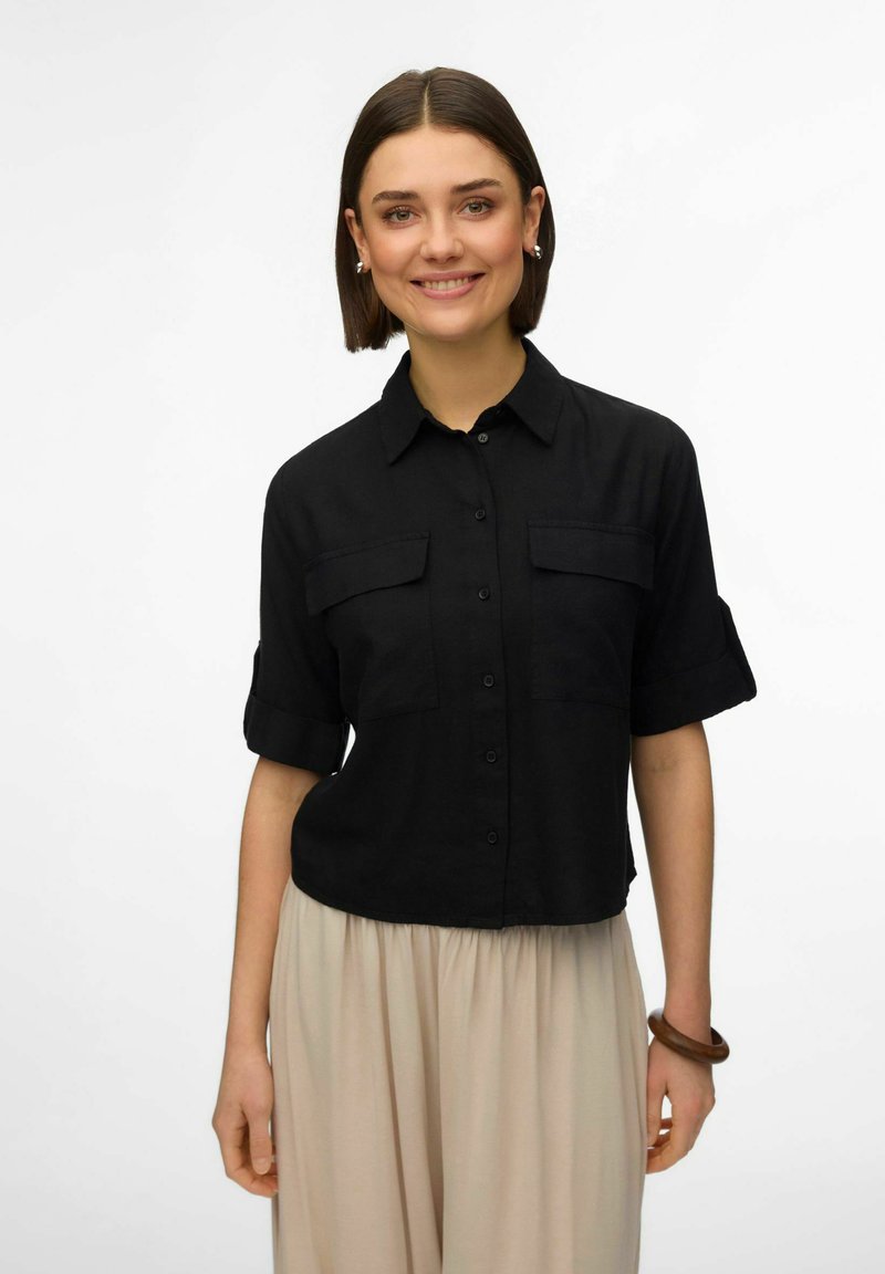 Femme aux cheveux bruns courts portant une chemise noire boutonnée avec les manches retroussées et un pantalon large beige, souriant devant un fond blanc.