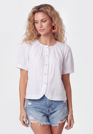 Frau mit lockigem blondem Haar, die eine weiße kurzärmlige Bluse mit Knöpfen, zerrissene Denim-Shorts und große bunte Creolen trägt, steht vor weißem Hintergrund.