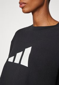 Černé bavlněné tričko s kulatým výstřihem, na přední straně velké bílé logo se třemi geometrickými tvary. Textura působí měkce.