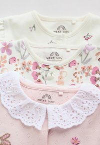 Kolme pastellisävyistä vauvan bodya pinottuna; kukkakuviot ja kirjotut eläimet, yhdessä valkea pitsikaulus, kaikki merkitty "NEXT baby."