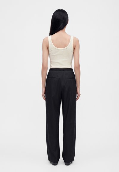 Femme aux longs cheveux noirs portant un haut blanc sans manches et un pantalon noir ample, debout, dos à un fond clair uni.
