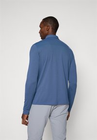 Majica z dolgimi rokavi in s kragnjo v enotni modri barvi, izdelana iz gladkega materiala. Ima diskreten logotip na levi rami.