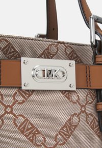 Sac à main à motif tan avec une texture tissée, des accents en cuir marron et un logo en métal sur une plaque métallique.