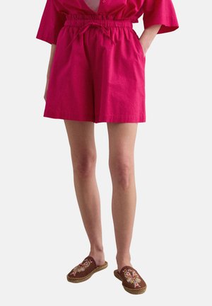 Mujer con pantalones cortos de talle alto y color rosa brillante con cordón y blusa de manga ancha, combinados con zapatos mocasín marrones bordados.