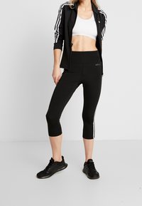 Černý sportovní outfit, který zahrnuje zkrácenou zipovou bundu s bílými pruhy a odpovídající legíny po kotníky. Textura je hladká, design je přiléhavý.