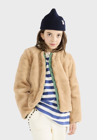 Faux fur beige jakke med en grøn trykknap-stolpe, båret over en blå og hvid stribet skjorte. Modellen har en marineblå strikket hat på.