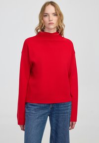 Woman with wavy blonde hair wearing a plain red long-sleeve sweater and blue jeans, standing against a light gray background.