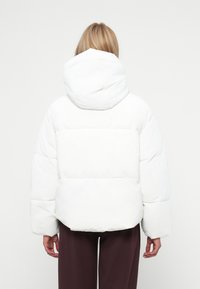 Jachetă albă din puf, cu glugă, având un design cusut, siluetă supradimensionată și textură moale. Pur și simplu peste pantaloni negri, largi.