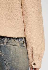 Beige struktureret sweater med en blød, fuzzy finish, der har en knapmanchet, parret med blå denimjeans med let falmning.