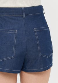Mørkeblå denimshorts med høy midje, baklommer og kontrastfarget hvit søm. Glatt tekstur med en skreddersydd passform.