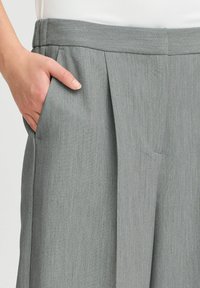 Grijze op maat gemaakte broek met een gladde textuur, plooien aan de voorkant en zijzakken, gepresenteerd met een hand in de zak.