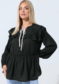 Nainen, jolla on pitkät vaaleat hiukset, pukeutuu mustaan pitkähihaiseen blouseen, jossa on röyhelöitä ja valkoinen solmu kaula-aukossa, yhdistettynä vaaleansinisiin farkkuihin.