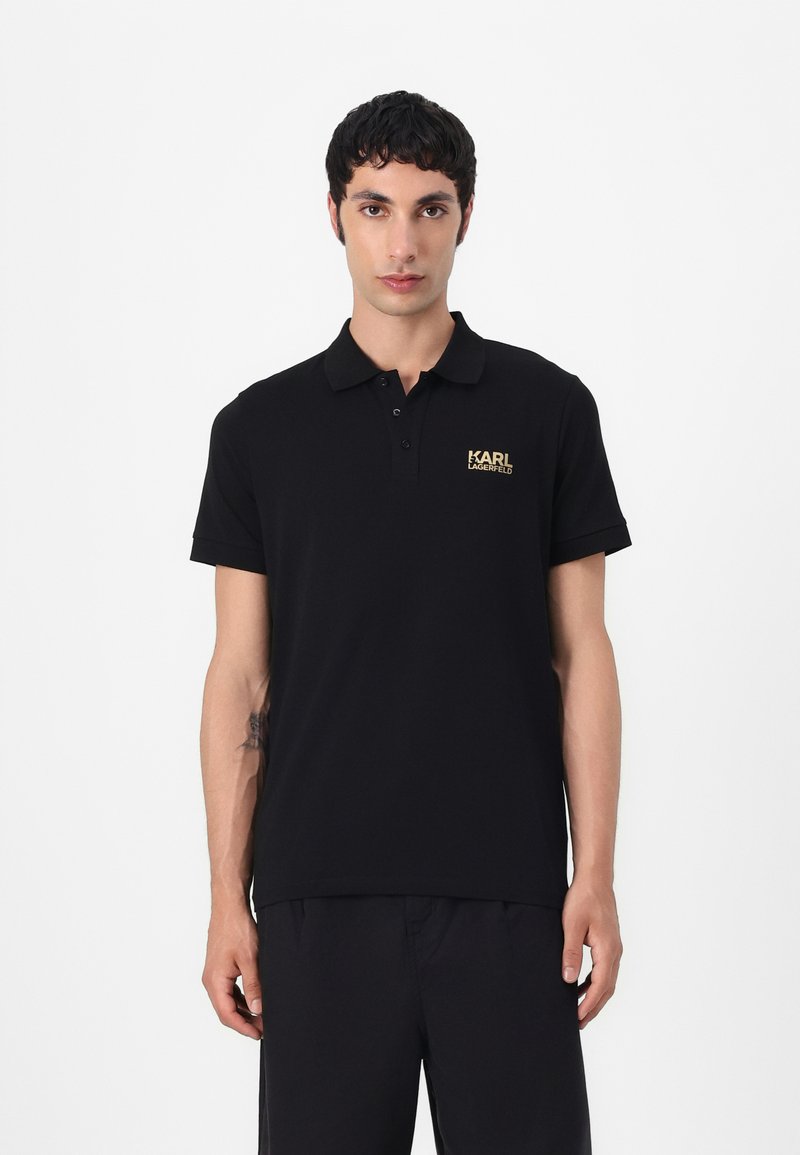 Zwarte polo met een kraag, korte mouwen en een gouden logo op de linkerborst. Gemaakt van gladde stof met een reguliere pasvorm.