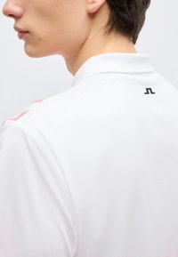 Πλάγια όψη ενός ατόμου που φοράει λευκό πουκάμισο με γιακά και μικρό μαύρο λογότυπο στο επάνω μέρος της πλάτης κοντά στον γιακά.