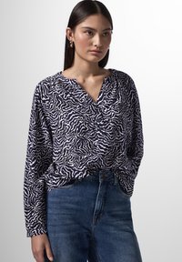 Marineblau und weiß gemusterte Bluse mit V-Ausschnitt, langen Ärmeln und lockerer Passform, kombiniert mit blauen Jeans. Der Stoff wirkt leicht und strukturiert.