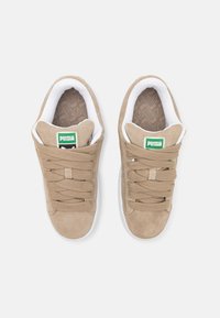 Baskets en daim beige avec semelles blanches, présentant des lacets épais et plats ainsi qu'un patch logo vert sur la languette. Motif texturé de la semelle intérieure visible.