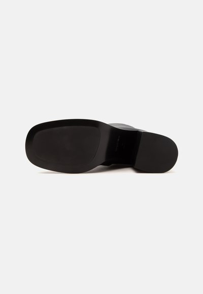 Botte en cuir noir vue par dessous avec une semelle lisse et un talon carré bas sur fond blanc.