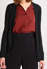 Cardigan negro con una textura moteada, llevado sobre una blusa burdeos con botones, combinado con pantalones negros de cintura alta. Diseño ajustado y casual.