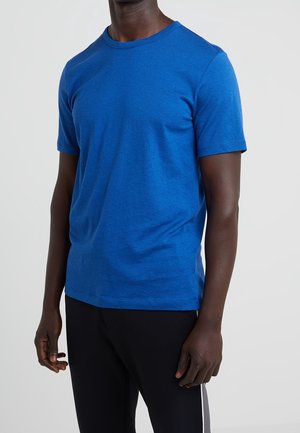 Mand iført en ensfarvet lys blå kortærmet t-shirt og sorte bukser mod en lysegrå baggrund.