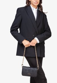 Bolso de mano negro acolchado con una correa de cadena dorada, con forma rectangular y logo V73. Estilizado con un blazer negro suelto y una camisa blanca.
