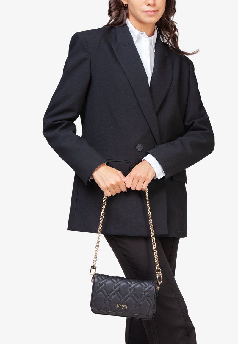 Bolso de mano negro acolchado con una correa de cadena dorada, con forma rectangular y logo V73. Estilizado con un blazer negro suelto y una camisa blanca.