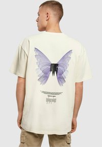 Camiseta de algodón color crema con un gran gráfico de mariposa morada en la parte de atrás. El diseño incluye texto negro y patrones intrincados.