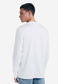 T-shirt bianco a maniche lunghe in cotone con scollatura rotonda, fessure laterali e vestibilità rilassata. Visualizzato dal retro.
