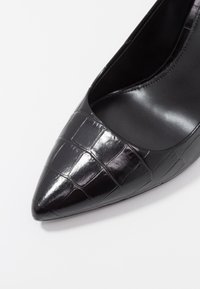 Chaussure en cuir verni noir à bout pointu avec une texture crocodile. Surface lisse avec un design épuré et un devant dégagé.
