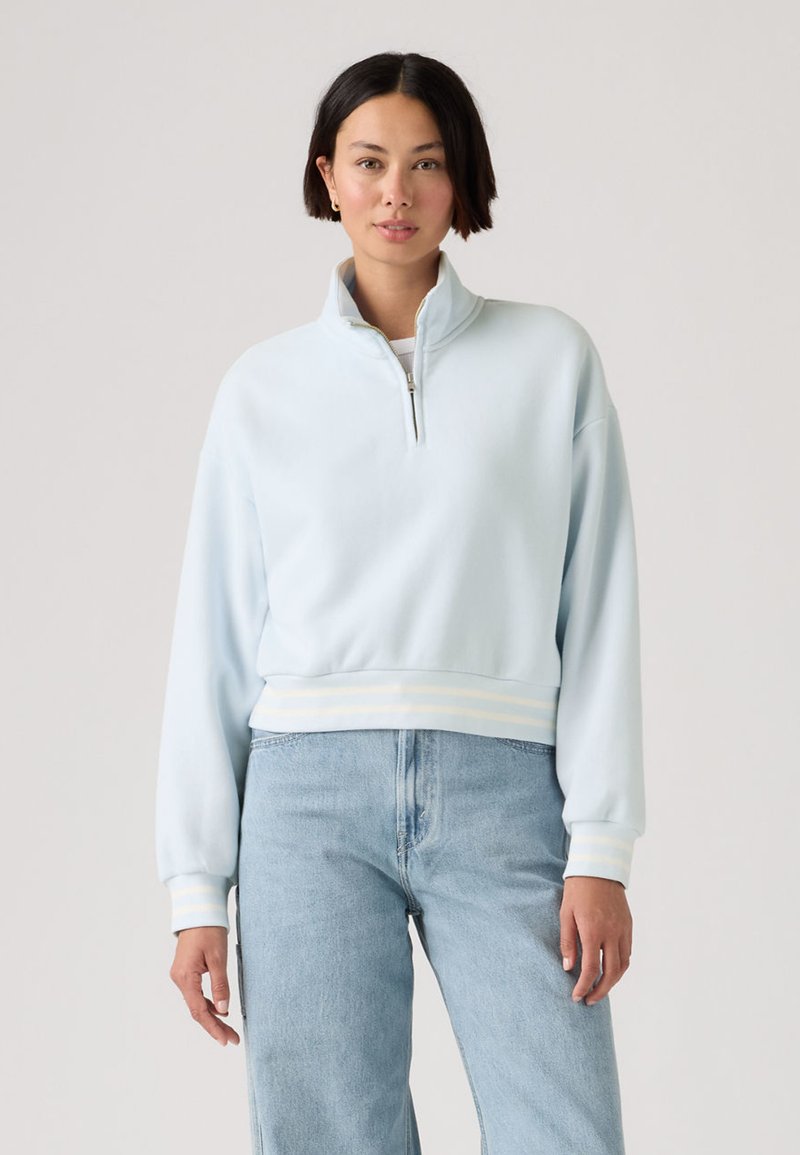 Femme aux cheveux courts et foncés portant un sweat-shirt à glissière quart de couleur bleu clair et un jean bleu clair, debout devant un fond uni.
