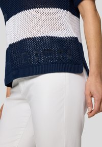 Gestreiftes Top in Navy und Weiß aus Strick mit offenem Webmuster, geripptem Rundhalsausschnitt und "GUESS" Logo-Akzent am Saum. Kombiniert mit weißen Hosen.