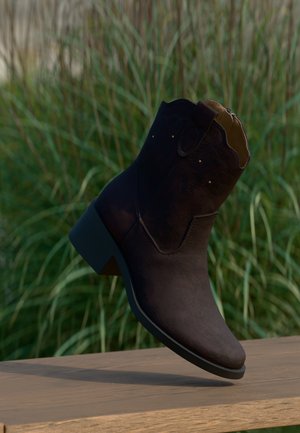 Bota al tobillo de ante marrón oscuro con tacón bajo, costuras decorativas y tiradores, exhibida sobre una superficie de madera con fondo de césped.