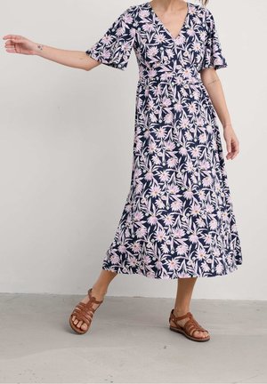 Vrouw die een marineblauwe midi-jurk met roze bloemenprint en bruine sandalen met bandjes draagt, staand met één arm uitgestrekt tegen een effen achtergrond.