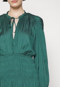 Femme portant un chemisier plissé de couleur sarcelle avec des manches froncées et un col à nouer, visible du cou à la taille sur un fond uni.