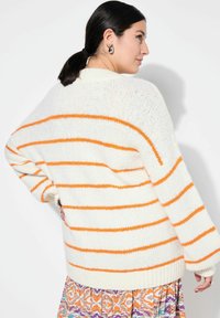 Weißer Pullover mit orangefarbenen Streifen, lockere Passform, gerippter Saum, strukturierter Strick, getragen über einem gemusterten Rock, mit weiten Ärmeln und überschnittenen Schultern.