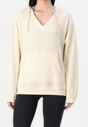 Mujer con sudadera con capucha de punto waffle color crema y bolsillo frontal, sobre una camiseta púrpura clara y leggings negros, mostrada desde los hombros hasta las caderas.