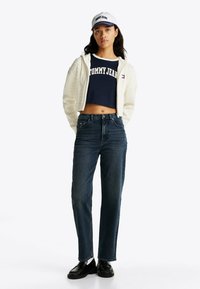 Chaquetón blanco acolchado con cremallera, camiseta corta azul marino con el logo "TOMMY JEANS", jeans rectos de mezclilla oscura, mocasines negros, gorra blanca.