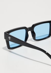 Ochelari de soare negri, unghiulari, cu lentile de nuanță albastră și brațe negre elegante. Detaliu logo notabil pe partea laterală a ramei.