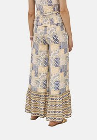 Pantalones acampanados y estampados en amarillo, azul y blanco, con diversos diseños florales y geométricos, y un dobladillo con volantes. Fabricados en tela ligera.