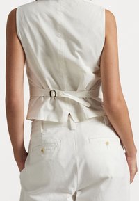 Bakvy av en person som bär en ärmlös vit topp med midjebälte och vita byxor med knäppta bakfickor.