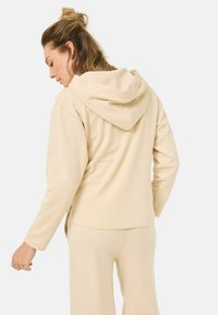 Beige hoodie met een dubbele capuchon, lange mouwen en zijsplitten. Gemaakt van zachte stof en gedragen met bijpassende beige broek.