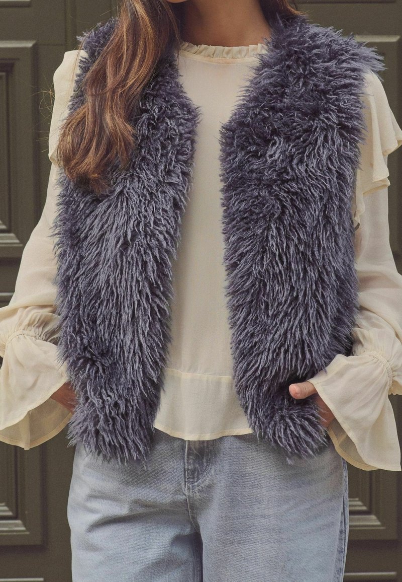 Femme portant un gilet en fausse fourrure gris sur une blouse crème à manches longues avec des poignets volantés et un jean bleu clair, mains dans les poches.