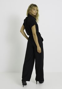 Mono negro con mangas cortas, cintura entallada y piernas anchas. Tela suave con un corte relajado. Combinado con tacones altos negros.