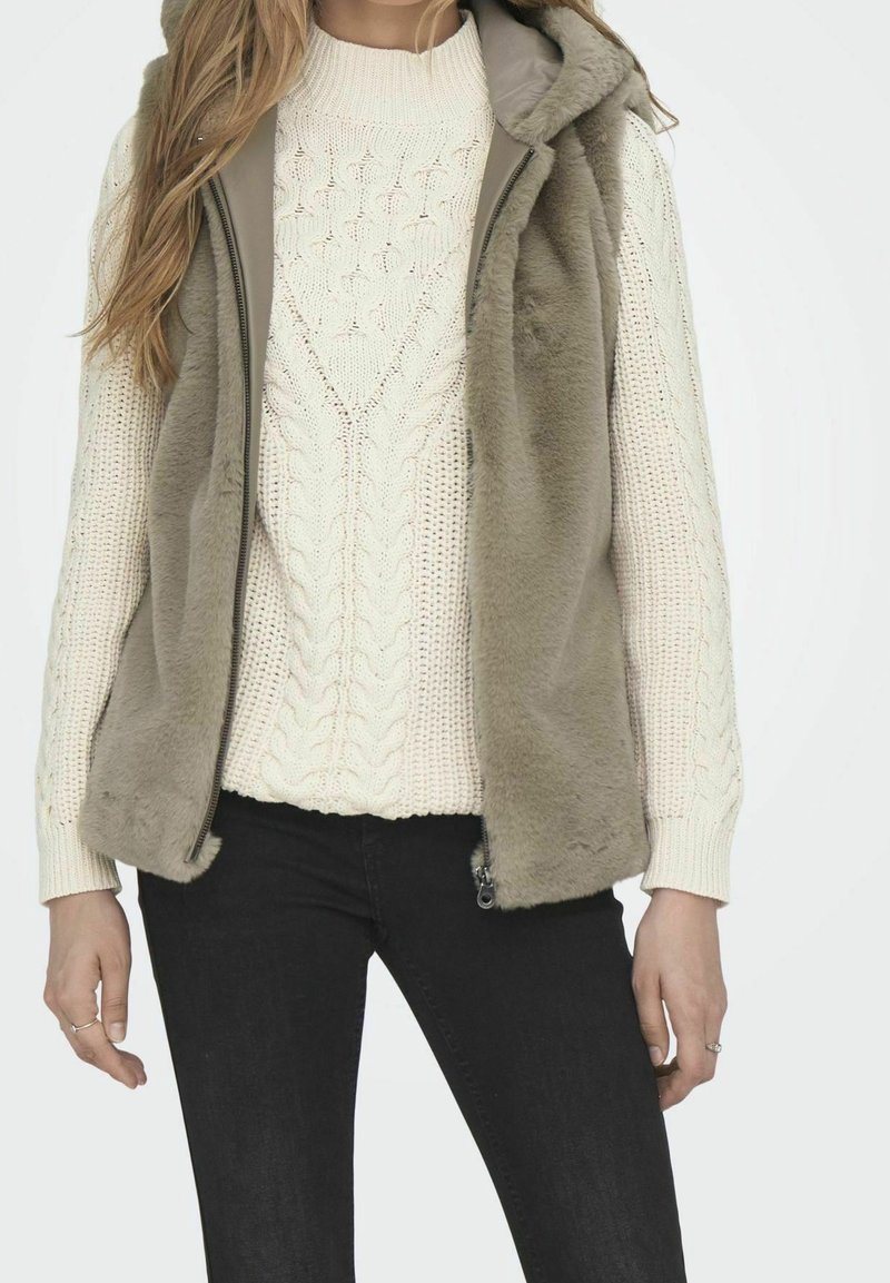Femme portant un pull crème en maille torsadée, un gilet sans manches zippé en fausse fourrure taupe et un pantalon noir sur un fond uni.
