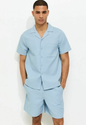 Młody mężczyzna stoi z rękami w kieszeniach, ubrany w jasnoniebieską koszulę z krótkim rękawem zapinaną na guziki i dopasowane szorty na jednolitym białym tle.