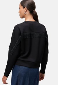 Černá mikina s třásněmi na ramenech a na zadní straně, strukturovaná tkanina, žebrované manžety a lem, nošená přes džínovou sukni.