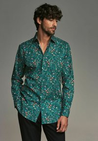 Cămașă cu model floral verde, cu guler deschis, mâneci lungi și hem curbat. Prezintă flori și frunze vibrante pe un material texturat.
