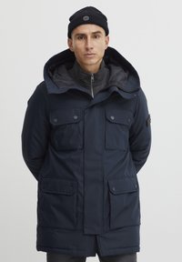 Marineblå isoleret parka med store frontlommer, hætte og lynlås foran. Har et blødt inderslag og en knapdetalje på ærmet.